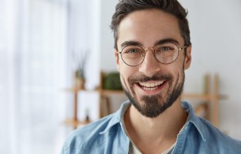 A smiling man with glasses and a beard wears a denim shirt. He stands in front of blurred shelves.